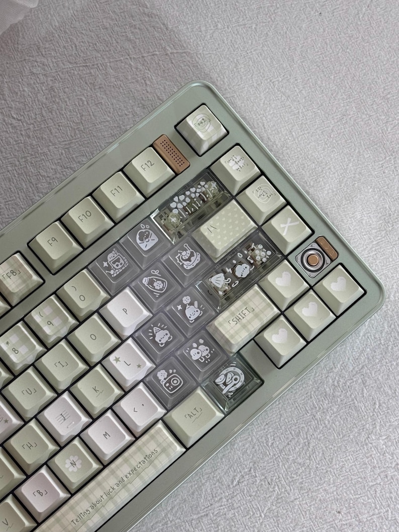 Puede incluir: Un teclado mec&aacute;nico personalizado con un marco verde claro y teclas blancas. Algunas teclas son transparentes con ilustraciones bonitas, mientras que otras tienen patrones de corazones y cuadros. El teclado tiene una tecla de acento marr&oacute;n y el texto "Telling about luck and expectations".