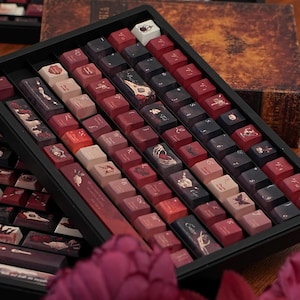 May include: A selection of custom keyboard keycaps displayed in a black tray. The keycaps showcase designs in red, black, and beige, with illustrations of skulls, butterflies, and gothic themes. The keycaps are arranged in rows, with a vintage book in the background.