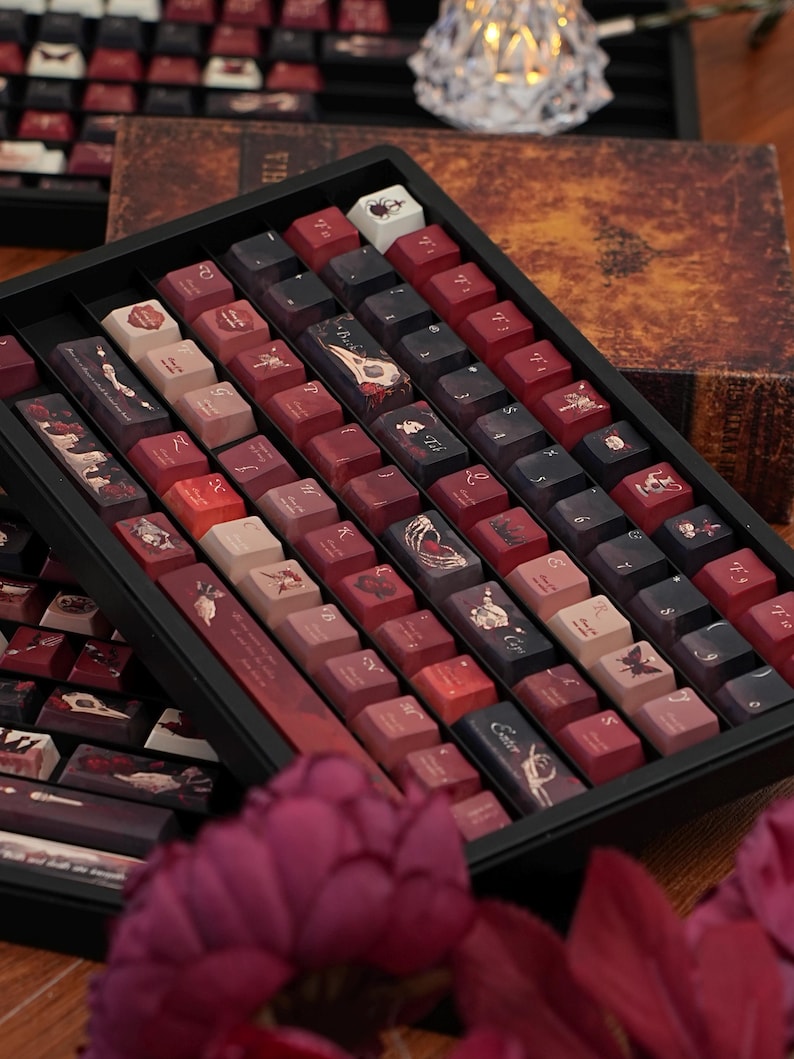 May include: A selection of custom keyboard keycaps displayed in a black tray. The keycaps showcase designs in red, black, and beige, with illustrations of skulls, butterflies, and gothic themes. The keycaps are arranged in rows, with a vintage book in the background.