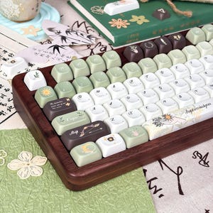 Puede incluir: Un teclado personalizado con marco de madera y una variedad de teclas. Las teclas son blancas, verdes y marrones, con varios s&iacute;mbolos y texto. El teclado est&aacute; sobre una superficie estampada con una taza y un libro en el fondo.