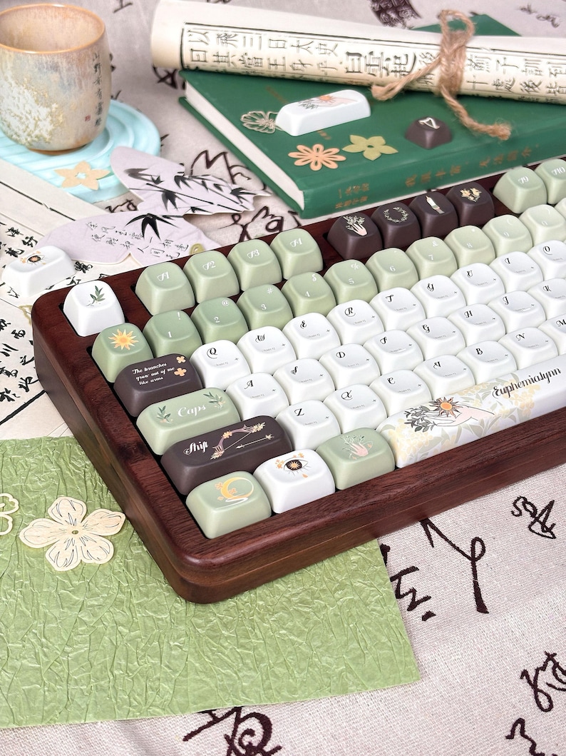 Puede incluir: Un teclado personalizado con marco de madera y una variedad de teclas. Las teclas son blancas, verdes y marrones, con varios s&iacute;mbolos y texto. El teclado est&aacute; sobre una superficie estampada con una taza y un libro en el fondo.