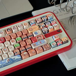 May include: A custom mechanical keyboard with a red frame and white keycaps. The keycaps feature colorful anime character designs. The keyboard is on a desk with a laptop and a magazine.