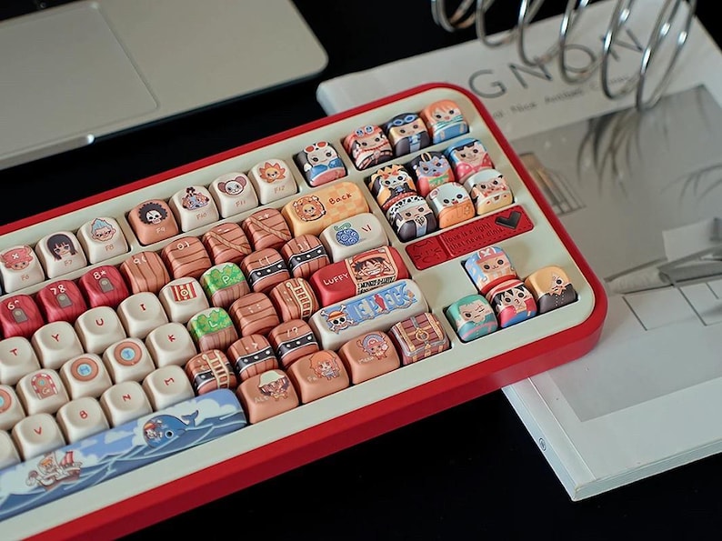 May include: A custom mechanical keyboard with a red frame and white keycaps. The keycaps feature colorful anime character designs. The keyboard is on a desk with a laptop and a magazine.