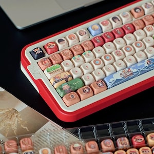 May include: A custom mechanical keyboard with a red and white frame. The keycaps feature colorful illustrations of anime characters. The keyboard is shown with a laptop in the background and a tray of extra keycaps.