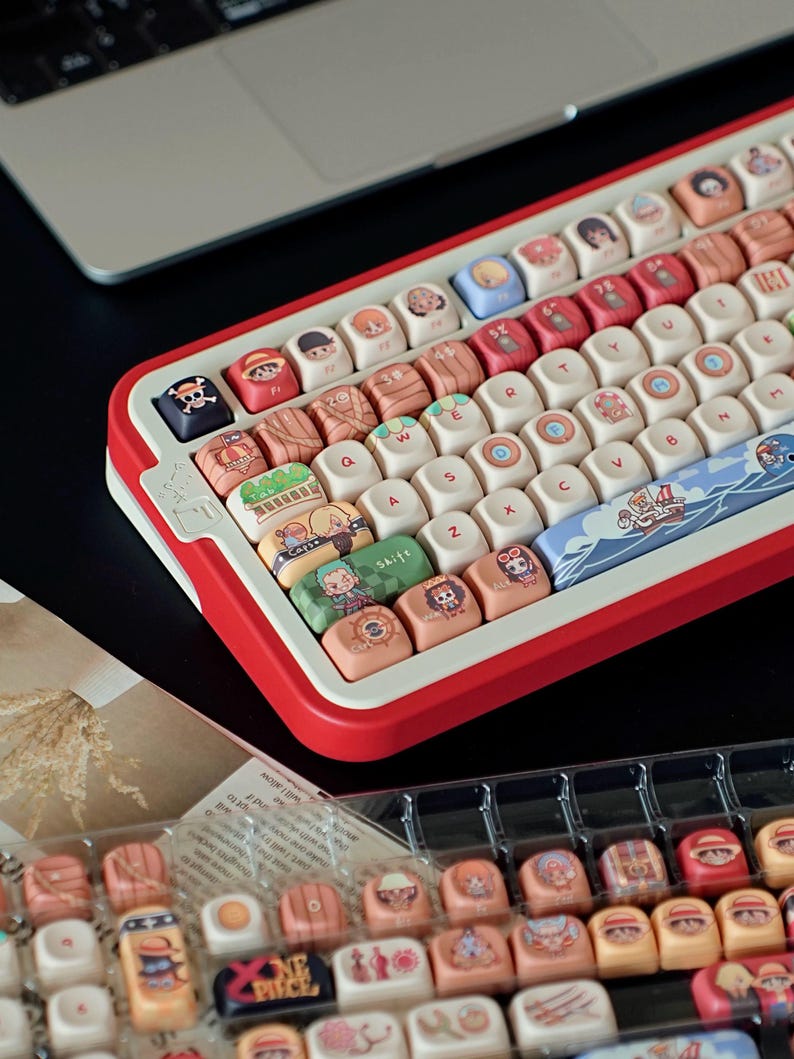May include: A custom mechanical keyboard with a red and white frame. The keycaps feature colorful illustrations of anime characters. The keyboard is shown with a laptop in the background and a tray of extra keycaps.