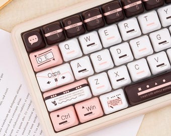 Puede incluir: Primer plano de un teclado de estilo retro con teclas blancas y rosas. Las teclas tienen una variedad de símbolos y texto, incluyendo "Caps", "Ctrl" y "Win". El teclado tiene un marco rosa claro y teclas marrones con la palabra "LODING".