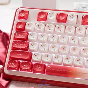 May include: A mechanical keyboard featuring a red and white color scheme with custom keycaps. The keys display various facial expressions, polka dots, and the text "The true beauty of life lies not in the destination but in the journey" on the spacebar.