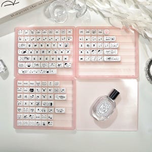 May include: A pink keyboard case with white keycaps decorated with cartoon animal designs. The case is open, showing the keyboard layout. A perfume bottle is in the lower right corner. The text "Rayon de soleil" is visible on a book.