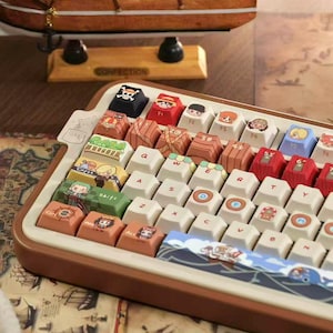 May include: A custom keyboard with keycaps featuring cartoon characters and nautical themes. The keyboard has a brown frame and white keys, with colorful illustrations on the keycaps. A wooden ship model sits in the background.