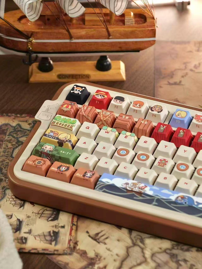 May include: A custom keyboard with keycaps featuring cartoon characters and nautical themes. The keyboard has a brown frame and white keys, with colorful illustrations on the keycaps. A wooden ship model sits in the background.