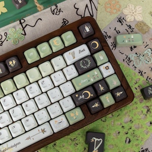 Puede incluir: Un teclado con marco de madera y teclas personalizadas en blanco, verde y marr&oacute;n. Las teclas presentan varios s&iacute;mbolos y dise&ntilde;os, incluyendo lunas, flores y texto. El teclado est&aacute; rodeado de papel decorativo y recortes florales.