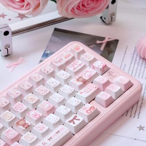 Może przedstawiać: Różowa klawiatura mechaniczna z białymi i różowymi nakładkami na klawisze. Nakładki na klawisze przedstawiają urocze ilustracje misiów, kokardek i innych wzorów. Klawiatura ma napis "BABY BEAR" na spacji. Różowe róże i inne elementy dekoracyjne są w tle.