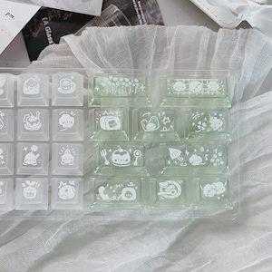 Puede incluir: Un conjunto de teclas de teclado cuadradas y transparentes con ilustraciones blancas de varios objetos y personajes. Algunas teclas est&aacute;n en un recipiente de pl&aacute;stico transparente, otras sueltas. Dise&ntilde;o lindo y caprichoso.