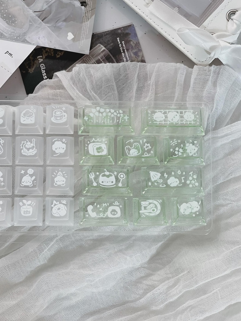 Puede incluir: Un conjunto de teclas de teclado cuadradas y transparentes con ilustraciones blancas de varios objetos y personajes. Algunas teclas est&aacute;n en un recipiente de pl&aacute;stico transparente, otras sueltas. Dise&ntilde;o lindo y caprichoso.