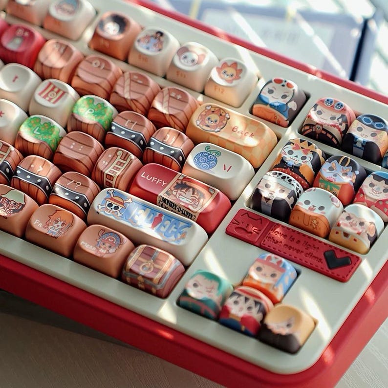 May include: A custom keyboard with keycaps featuring colorful anime character designs. The keycaps have various illustrations and text, including "LUFFY" and "MONKEY-D-LUFFY". The keyboard has a red frame and a light-colored base.