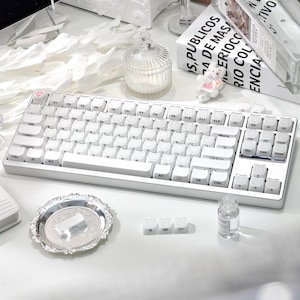 May include: A white mechanical keyboard with a compact layout, featuring white keycaps and a pink paw print on the escape key. The keyboard is on a white surface, with a decorative tray and small glass jar nearby. The image also includes a book with text.