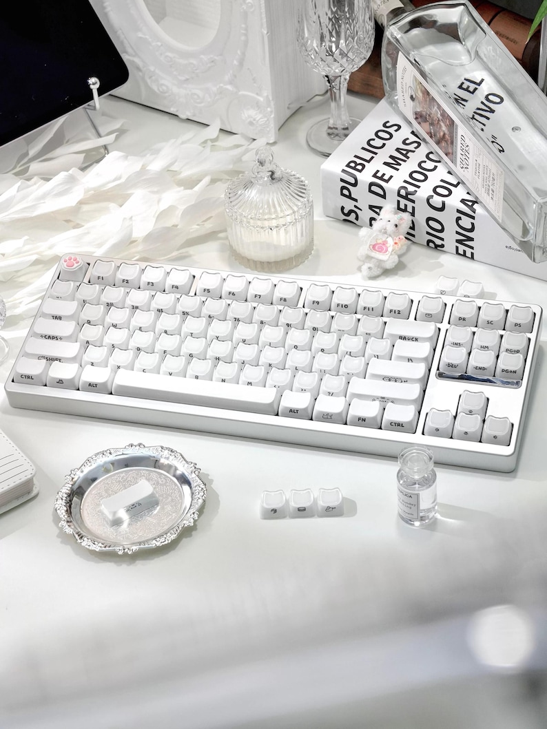 May include: A white mechanical keyboard with a compact layout, featuring white keycaps and a pink paw print on the escape key. The keyboard is on a white surface, with a decorative tray and small glass jar nearby. The image also includes a book with text.