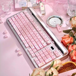 May include: A pink and silver computer keyboard with pink keycaps. The keycaps have white lettering and cute animal illustrations. The keyboard is surrounded by pink accessories and flowers.