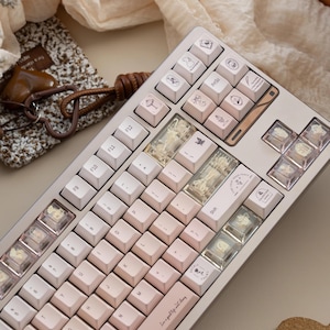 May include: A light pink mechanical keyboard with white and pink keycaps. Some keycaps have small illustrations. The keyboard has a copper-colored accent and the text "Live a good life with love" is printed on the space bar.