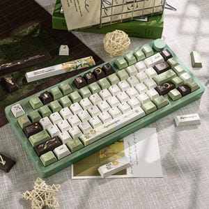 Puede incluir: Un teclado mec&aacute;nico verde y blanco con teclas personalizadas. Las teclas presentan dise&ntilde;os florales y celestiales, con algunas teclas en tonos marrones y verdes. El teclado est&aacute; rodeado de elementos decorativos, incluyendo una peque&ntilde;a estrella y una tarjeta con texto.