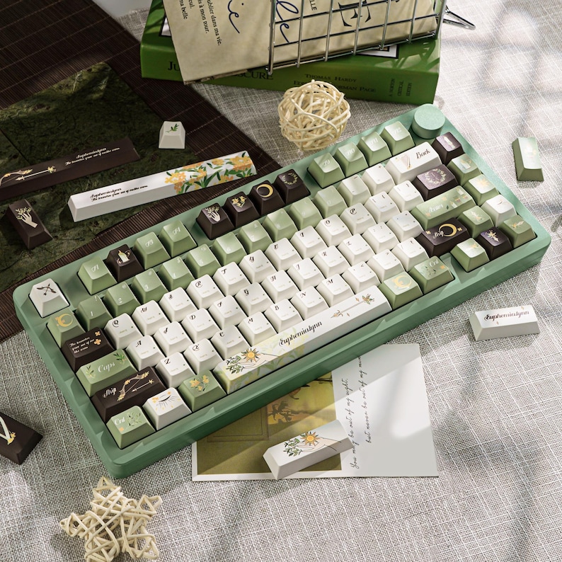 Puede incluir: Un teclado mec&aacute;nico verde y blanco con teclas personalizadas. Las teclas presentan dise&ntilde;os florales y celestiales, con algunas teclas en tonos marrones y verdes. El teclado est&aacute; rodeado de elementos decorativos, incluyendo una peque&ntilde;a estrella y una tarjeta con texto.