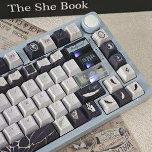 May include: A light blue mechanical keyboard with custom keycaps. The keycaps are a mix of white and dark grey, featuring various designs like feathers, butterflies, and other graphics. The keyboard has a volume knob and the text "Background" is visible on one of the keys.