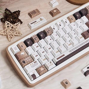 May include: A beige and white mechanical keyboard with custom keycaps. The keycaps feature a mix of white, brown plaid, and coffee-themed designs. Some keys have numbers, symbols, and coffee cup icons. The keyboard has a brown border.