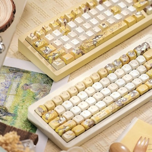 Könnte beinhalten: Zwei mechanische Tastaturen mit gelben und weißen Tastenkappen. Die Tastenkappen zeigen Cartoon-Charakter-Designs in Gelb und Braun. Die Tastaturrahmen sind hellgelb. Die Tastaturen befinden sich auf einer hellen Oberfläche.