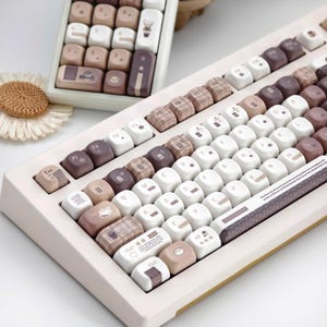 Puede incluir: Un teclado mecánico personalizado con un marco color crema y teclas en tonos blanco, marrón y morado. Algunas teclas presentan patrones e iconos decorativos. Un juego de teclas separado es visible en el fondo.