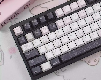 May include: A black mechanical keyboard with white and dark gray keycaps. The keycaps have various symbols and text, including numbers, letters, and special characters. The keyboard is angled towards the viewer, with a pink book visible in the background.