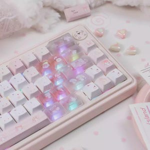May include: A pastel pink mechanical keyboard with white and translucent keycaps. The keycaps feature cartoon character designs. The keyboard has a light-up feature, and the keycaps are arranged on a pink base. Decorative heart-shaped beads are scattered around.