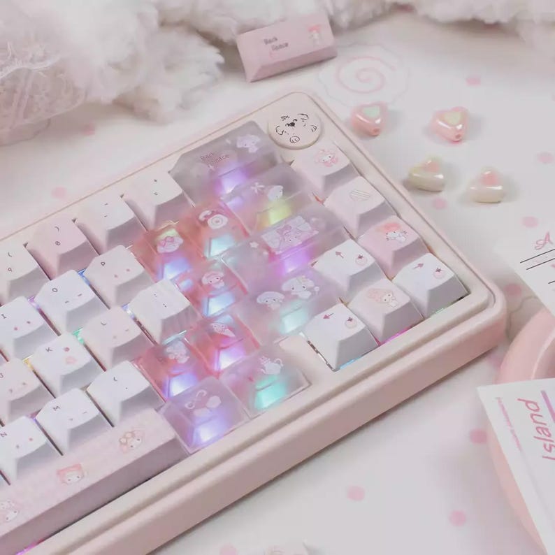May include: A pastel pink mechanical keyboard with white and translucent keycaps. The keycaps feature cartoon character designs. The keyboard has a light-up feature, and the keycaps are arranged on a pink base. Decorative heart-shaped beads are scattered around.