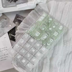 Puede incluir: Un recipiente de pl&aacute;stico transparente que contiene un juego de teclas de teclado verde claro y blanco. Cada tecla presenta una ilustraci&oacute;n blanca &uacute;nica. Las teclas est&aacute;n dispuestas sobre una superficie blanca texturizada, con un fondo borroso.