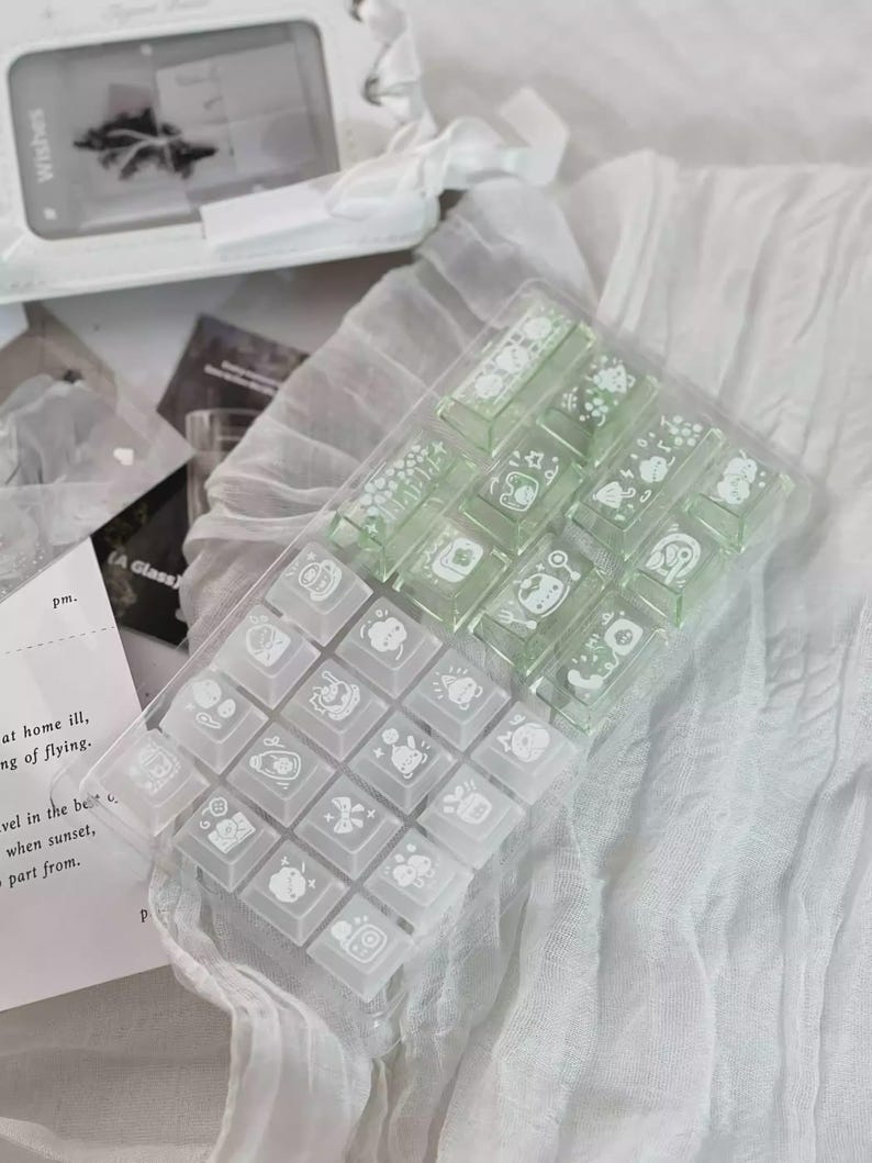 Puede incluir: Un recipiente de pl&aacute;stico transparente que contiene un juego de teclas de teclado verde claro y blanco. Cada tecla presenta una ilustraci&oacute;n blanca &uacute;nica. Las teclas est&aacute;n dispuestas sobre una superficie blanca texturizada, con un fondo borroso.