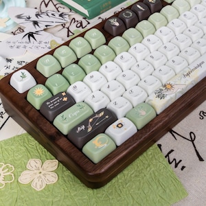 Puede incluir: Un teclado mec&aacute;nico personalizado con un marco de madera y una variedad de teclas en blanco, verde y marr&oacute;n. Algunas teclas presentan dise&ntilde;os bot&aacute;nicos y celestiales, y texto. El teclado est&aacute; en &aacute;ngulo, mostrando las teclas en detalle.