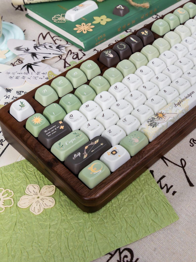 Puede incluir: Un teclado mec&aacute;nico personalizado con un marco de madera y una variedad de teclas en blanco, verde y marr&oacute;n. Algunas teclas presentan dise&ntilde;os bot&aacute;nicos y celestiales, y texto. El teclado est&aacute; en &aacute;ngulo, mostrando las teclas en detalle.