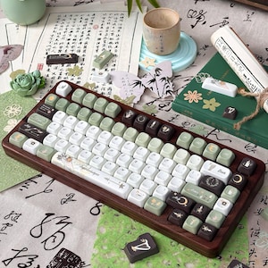 Puede incluir: Un teclado mec&aacute;nico personalizado con marco de madera y teclas en tonos verdes, blancos y marrones. Las teclas presentan dise&ntilde;os florales y celestiales. El teclado est&aacute; rodeado de objetos decorativos, incluyendo una taza y un libro.