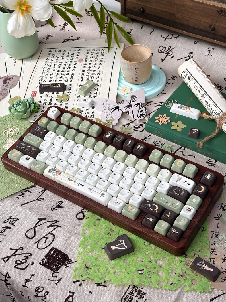 Puede incluir: Un teclado mec&aacute;nico personalizado con marco de madera y teclas en tonos verdes, blancos y marrones. Las teclas presentan dise&ntilde;os florales y celestiales. El teclado est&aacute; rodeado de objetos decorativos, incluyendo una taza y un libro.