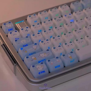 Pode incluir: Um teclado mecânico personalizado com teclas brancas e azul claro. As teclas apresentam desenhos e ícones de gatos de desenhos animados. O teclado tem uma estrutura prateada e teclas iluminadas. As teclas têm texto como "ESC", "TAB", "CAPS", "SHIFT", "CTRL", "WIN" e "ALT".
