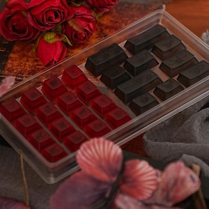 May include: A transparent plastic container holding red and black keycaps. The red keycaps are in the foreground, and the black keycaps are behind them. Red roses are in the background, with a blurred flower in the foreground. The keycaps are likely for a keyboard.