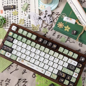 Puede incluir: Un teclado con marco de madera con teclas blancas, verdes y negras. Las teclas presentan varios dise&ntilde;os, incluyendo motivos florales y celestiales, y la palabra "Euphemiakynn". El teclado est&aacute; rodeado de papel decorativo, una taza y otros art&iacute;culos de papeler&iacute;a.