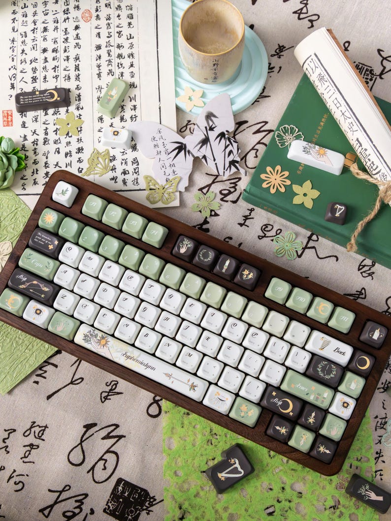 Puede incluir: Un teclado con marco de madera con teclas blancas, verdes y negras. Las teclas presentan varios dise&ntilde;os, incluyendo motivos florales y celestiales, y la palabra "Euphemiakynn". El teclado est&aacute; rodeado de papel decorativo, una taza y otros art&iacute;culos de papeler&iacute;a.