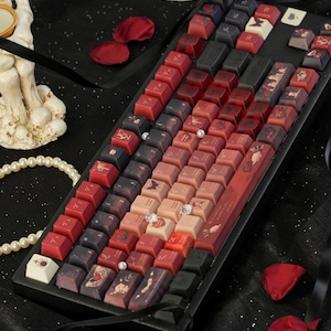 May include: A custom mechanical keyboard with keycaps in shades of red, black, and cream. The keycaps feature various designs, including floral patterns and illustrations. The keyboard sits on a black surface, with rose petals and a pearl necklace nearby.