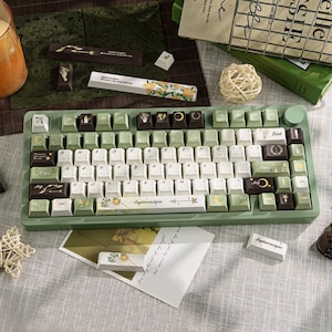 Puede incluir: Un teclado mec&aacute;nico verde con teclas blancas, verdes y marrones. Las teclas presentan dise&ntilde;os florales y celestiales. El teclado tiene un marco verde a juego y una rueda giratoria. El texto "Ephermiskyn" est&aacute; impreso en una tecla.