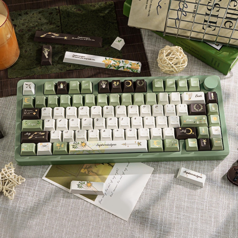 Puede incluir: Un teclado mec&aacute;nico verde con teclas blancas, verdes y marrones. Las teclas presentan dise&ntilde;os florales y celestiales. El teclado tiene un marco verde a juego y una rueda giratoria. El texto "Ephermiskyn" est&aacute; impreso en una tecla.