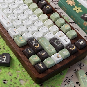 Puede incluir: Un teclado mec&aacute;nico personalizado con teclas blancas, verdes y marrones. Las teclas presentan varios dise&ntilde;os bot&aacute;nicos y celestiales. El teclado est&aacute; en un marco de madera, con un libro verde y papel decorativo en el fondo.