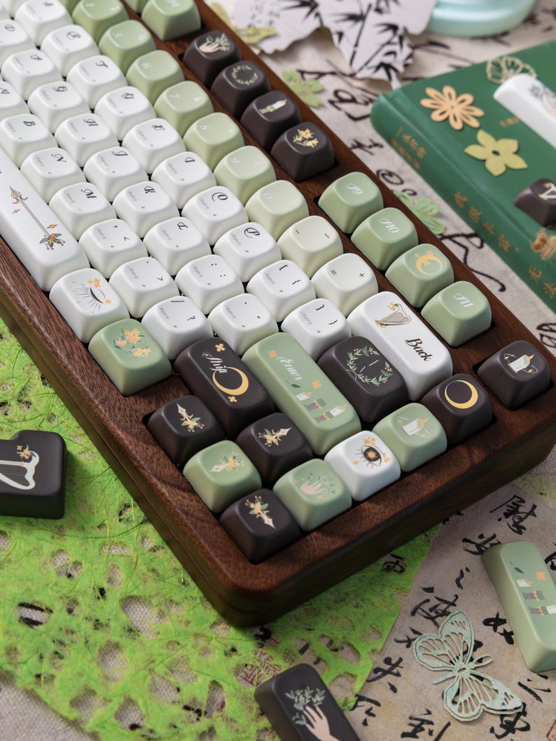 Puede incluir: Un teclado mec&aacute;nico personalizado con teclas blancas, verdes y marrones. Las teclas presentan varios dise&ntilde;os bot&aacute;nicos y celestiales. El teclado est&aacute; en un marco de madera, con un libro verde y papel decorativo en el fondo.