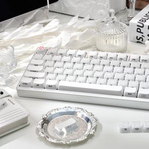 May include: A white mechanical keyboard with white keycaps, a silver decorative tray, and a small glass jar with a lid. The keyboard has a paw print keycap. Other items include a small notebook and a decorative frame.