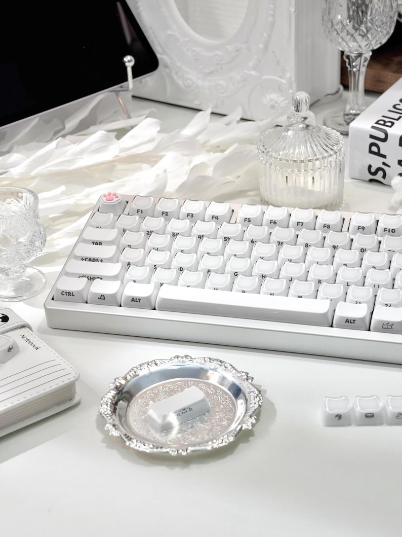 May include: A white mechanical keyboard with white keycaps, a silver decorative tray, and a small glass jar with a lid. The keyboard has a paw print keycap. Other items include a small notebook and a decorative frame.