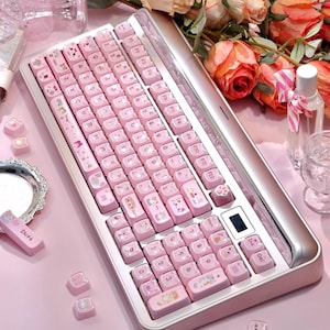 May include: A pink mechanical keyboard with cat-themed keycaps and a silver frame. The keyboard has a small digital display. Additional keycaps are scattered on a pink surface. The text "CAT BUTTERFLY" is visible.